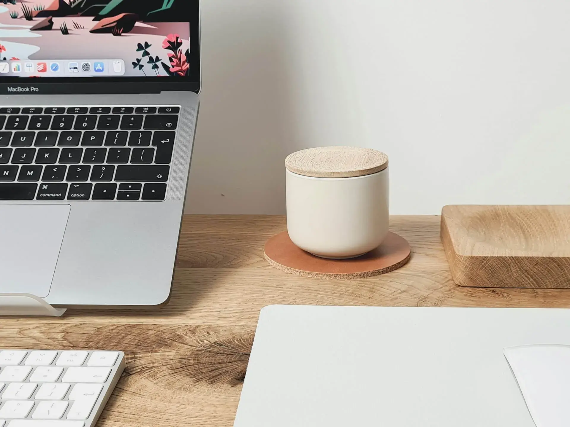 Open MacBook Pro on a wooden desk with a flat-illustrated landscape wallpaper, a white ceramic candle with cork lid on a leather coaster, and a white Magic Keyboard in the foreground