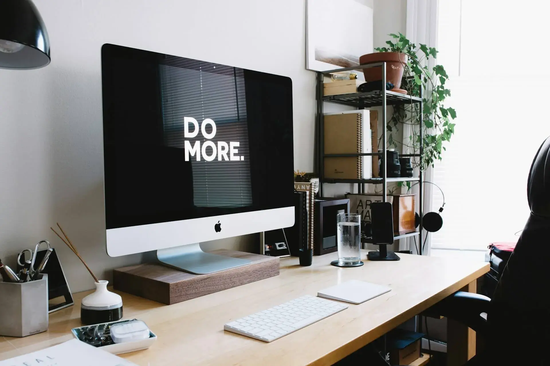 Apple iMac in a bright minimalist home office showing a black-and-white 'DO MORE' wallpaper, on a wooden desk beside a wire shelving unit with notebooks and a potted ivy plant